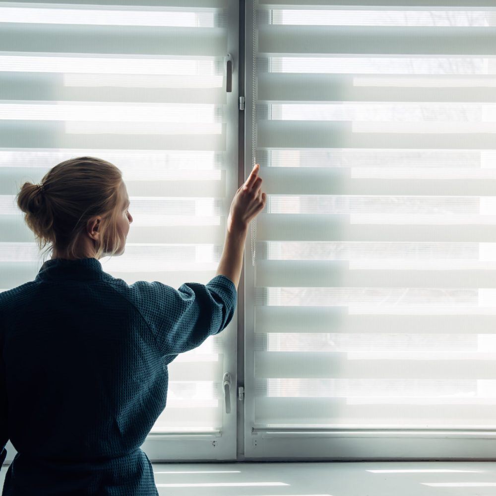 Woman's silhouette at window in the background light. Young woman in gown stands near window with blinds. Quarantine, self-isolation, stay home, self-preservation, coronavirus pandemic.
